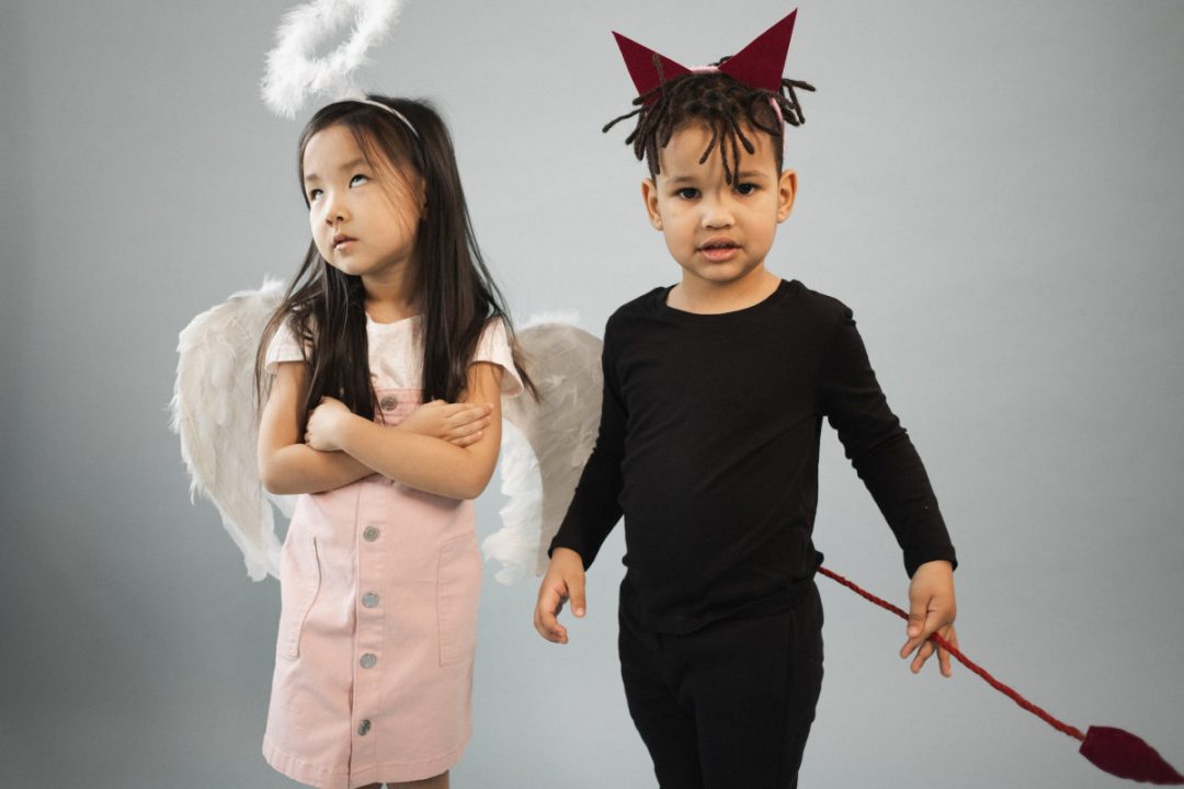 Adorable Asian girl in angel costume with halo standing near black boy in devil outfit looking at camera on gray background in studio